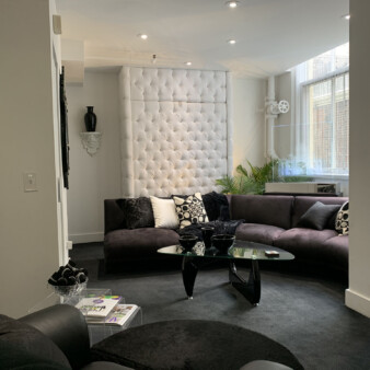 Modern living room in a studio apartment with high ceilings and industrial elements, decorated in black and white with shades of grey. Sectional sofa in grey with various pillows. Glass coffee table with black base and black glass bowls decorating it. All walls are white, with a couple of accent details on the walls. Laser etched glass sculpture of the Taj Mahal on the right side of the room, with large window behind. On the right side there are two white columns with three glass pieces in between serving as serve shelves.