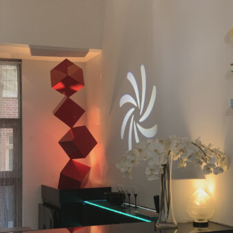 Modern decoration of the kitchen space with a sculpture made of red vertically aligned three-dimensional boxes attached to one another from the tip of one corner placed on top of black refrigerator. There is also a projection of a six elongated petal-like shapes in white light on the wall. There are other accents in black, including tall glasses and vases.