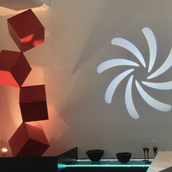 Image of kitchen by a window facing the outside. It shows a sculpture made of red vertically aligned three-dimensional boxes attached to one another from the tip of one corner and of a projection of a six elongated petal-like shapes in white light on the wall.