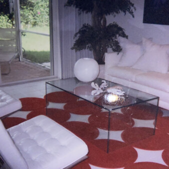 Living room with a white couch with white pillows and a rectangular glass coffee table with a spherical lamp on top. The table is sitting on a red and white carpet with a diamond pattern. Across from the table are two white designer chairs. A tree sits in the corner of the room. A glass sliding door open up to the back yard.