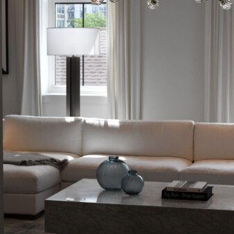 Spacious living room with tall ceiling, sectional sofa in white fabric, long white curtains on the side of windows hanging from ceiling to floor. Living room table is a rectangular piece in grey and white with two decorative glass bowls. A couple of modern lamps adorn the space, one behind the sofa and one hanging from the ceiling over the dining room table.