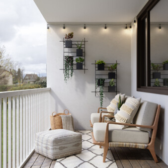 Balcony decorated with two chairs with simple lines with off-white fabric, complemented with two large pillows of the same color but with textured details. On a wall by the chairs there are two metal structures made of thin, black metal bars that hold black rectangular container with texturized surface for different plants. On the floor in front of the chairs there is an off-white pouf with geometrical designs in sand color.