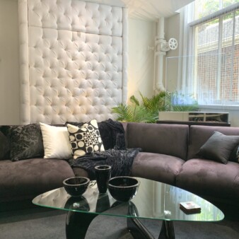 Coffee table with a grey couch behind it and a plush wall treatment in the background photograph of interior featuring a glass coffee table with a grey couch behind it and a plush wall treatment in the background
